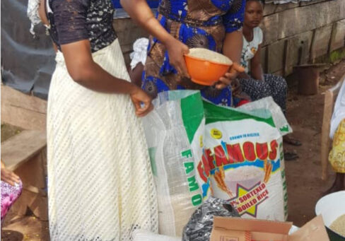 This is an image of two women standing over a large bag of what looks like rice or some sort of grain. The woman on the left is wearing a long white skirt and her hair is wrapped in white fabric that drapes down her neck. The other woman is leaning over the bag with an orange bowl. She is wearing a maroon head wrap and patterned blue and gold dress. In the foreground of the photo you can see additional bags and boxes of food.