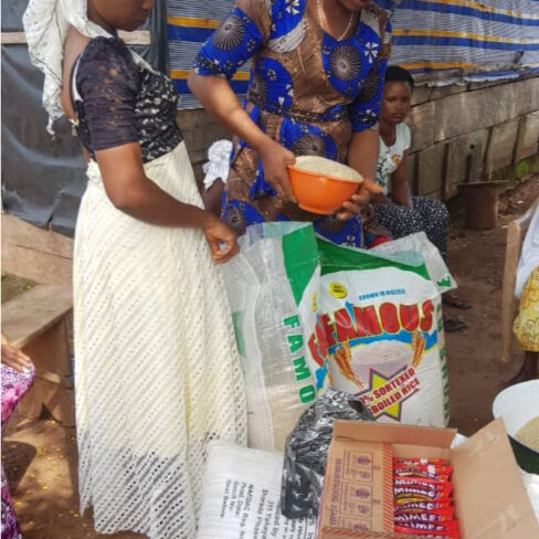 This is an image of two women standing over a large bag of what looks like rice or some sort of grain. The woman on the left is wearing a long white skirt and her hair is wrapped in white fabric that drapes down her neck. The other woman is leaning over the bag with an orange bowl. She is wearing a maroon head wrap and patterned blue and gold dress. In the foreground of the photo you can see additional bags and boxes of food.