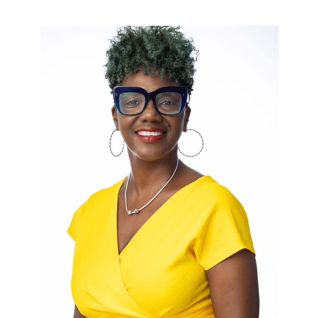 michellecropped michelle portrait. A woman with short curly salt and pepper hair. She's smiling confidently for a portrait shot and is wearing a bright solid short-sleeved yellow dress. She is also wearing expressive large blueish purple glasses, hoop earrings, and a necklace