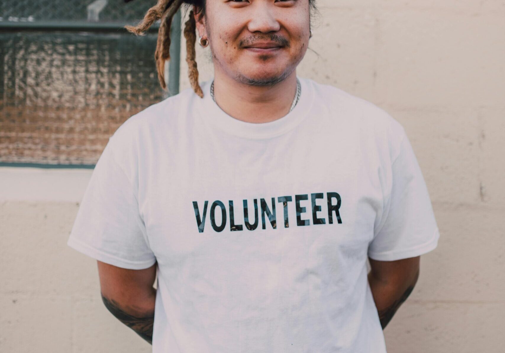 This is a photo of a young man with dreadlocs that are dark at the roots and more blonde towards the end. He's standing with his hands behind his back and he's smiling in a very warm, friendly way. His shirt is white and clearly says "Volunteer" on the front. He's standing in front of a white concrete wall with a window.